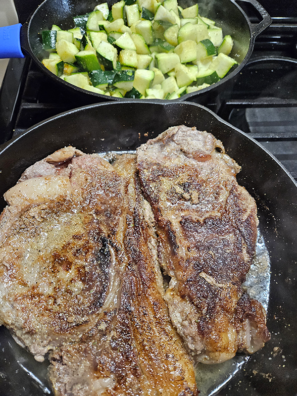 Two cast iron skillets, one with steak and one with zucchini cooking on a stove
