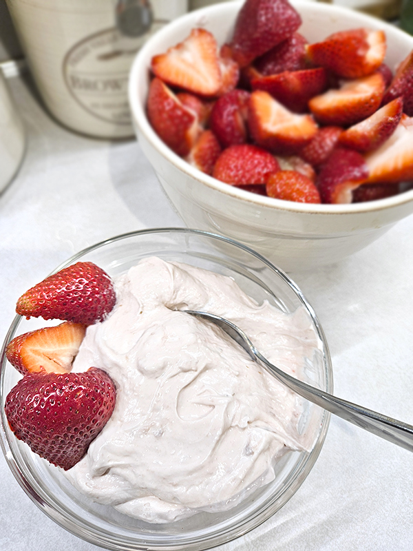 A bowl of marshmallow strawberry fruit dip in front of a bowl of halved strawberries