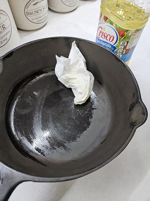 A cast iron skillet getting oil wiped on it with a paper towel.