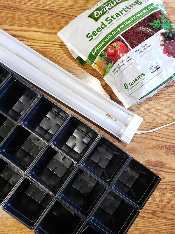For Part 1, a seed tray, bag of seed starting soil, and a led shop light sitting on a wooden floor are the main supplies you need to start seeds indoors.  