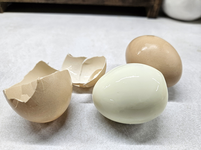 A hard boiled egg out of the shell with the shell next to it and one egg still in the shell. 